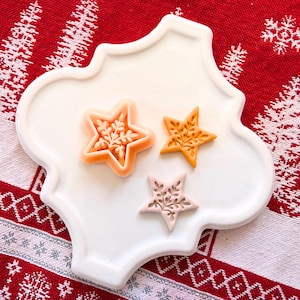 May include: Three star-shaped decorative soaps with floral designs rest on a white, scalloped-edge plate. The soaps are in shades of orange and pink, set against a red and white patterned background, suggesting a holiday theme.