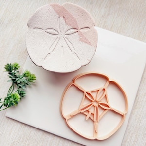 Può includere: Un set di due stampini per biscotti a forma di dollaro di sabbia. Lo stampino più grande è in ceramica bianca con una glassa rosa e un design a dollaro di sabbia in rilievo. Lo stampino più piccolo è in plastica marrone chiaro e presenta un design a dollaro di sabbia dettagliato.