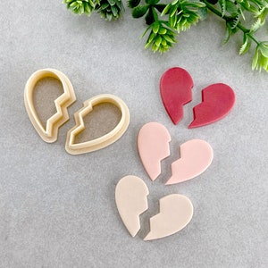 May include: A set of four broken heart cookie cutters and fondant decorations. The cutters are made of metal and are in the shape of a heart that has been broken in half. The fondant decorations are made of sugar paste and are in the colors red, pink, and white.