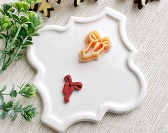 May include: Two polymer clay ornaments in the shape of bows. One bow is red and the other is a light peach color. The ornaments are on a white ceramic plate with a scalloped edge.