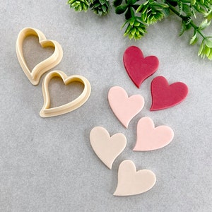以下が含まれることがあります： ハート型のクッキーカッター2つと、ピンクと赤の色合いのハート型のフォンダン装飾6つ。フォンダン装飾は1列に並べられており、クッキーカッターは画像の左側にある。