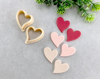 Puede incluir: Dos cortadores de galletas en forma de corazón y seis decoraciones de fondant en forma de corazón en tonos de rosa y rojo. Las decoraciones de fondant están dispuestas en una fila, con los cortadores de galletas en el lado izquierdo de la imagen.