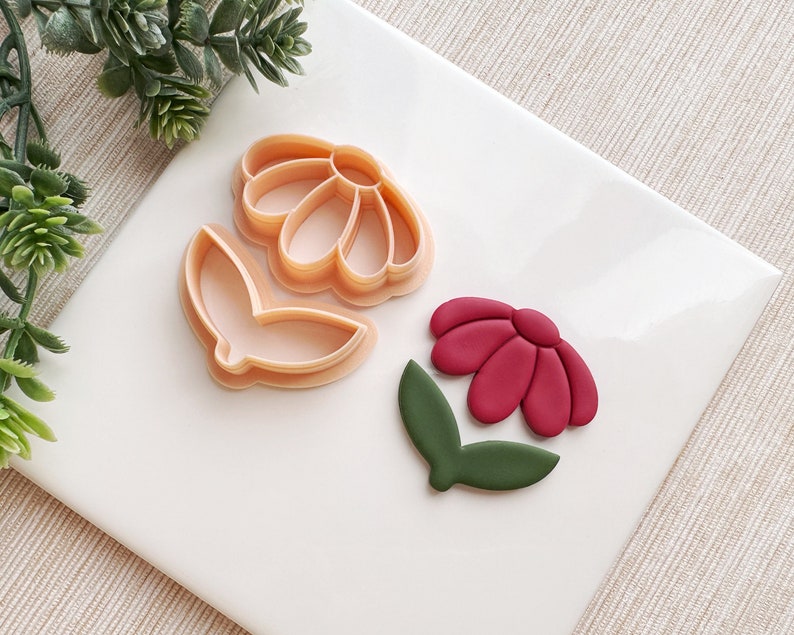 May include: Two plastic cookie cutters in the shape of a flower, one with a leaf. A red flower with a green leaf is made from polymer clay.