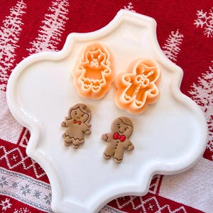 Peut inclure: Une assiette blanche décorative en forme de quadrilobe contient deux emporte-pièces en pain d'épices et deux biscuits en pain d'épices. Les biscuits sont bruns avec des nœuds papillon rouges. Les emporte-pièces sont orange.