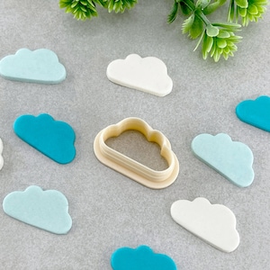 May include: A set of nine cloud-shaped cookie cutters, including one larger cutter with a raised edge. The cutters are made of white plastic and are arranged on a grey surface. There are also several blue and light blue fondant clouds.