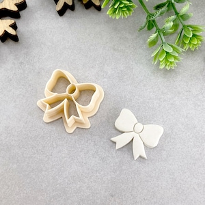 May include: Two plastic cookie cutters in the shape of bows. The larger bow is a double layer and the smaller bow is a single layer.
