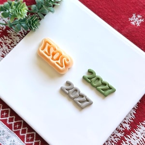 May include: A peach-colored plastic cookie cutter shaped like the number 2021, accompanied by two small clay or fondant cutouts of the number 2021 in sage green and light gray, all arranged on a smooth white surface.