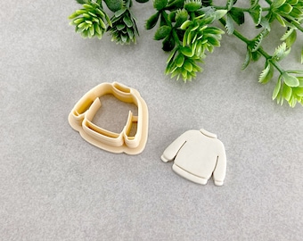 Puede incluir: Dos cortadores de galletas con forma de suéter. Los cortadores están hechos de plástico blanco y están sobre una superficie gris.