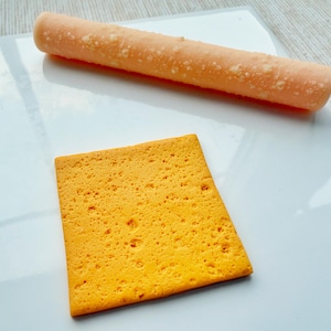 May include: A close-up of a yellow square cracker with a textured surface. A pink and white popsicle is also in the image.