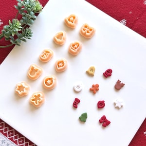 Puede incluir: Una colección de cortadores de galletas de plástico color melocotón con diversas formas festivas junto a pequeños adornos de arcilla terminados en rojo, verde, blanco, marrón y naranja, con diseños de hombrecitos de jengibre, calcetines y lazos.