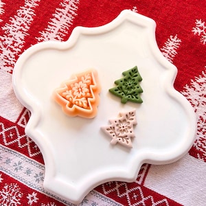 以下が含まれることがあります： 桃色、緑色、ピンク色のクリスマスツリー型の装飾品が3つ。それぞれ雪の結晶のデザインが施されています。白い縁の波打つお皿の上に配置され、赤と白の模様の背景に置かれています。