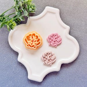 Może przedstawiać: Trzy wykrawacze do ciastek w kształcie muszli w odcieniach pomarańczu i różu spoczywają na białym talerzu z falistą krawędzią. Wykrawacze mają wzory kwiatowe i liściaste. Gałązka zielonych liści znajduje się w lewym górnym rogu.