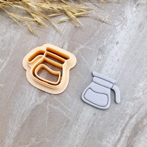 May include: Two coffee pot-shaped cookie cutters. One is a light tan plastic cutter, and the other is a grey, finished cookie. The cutters are on a grey marble surface with dried wheat stalks.