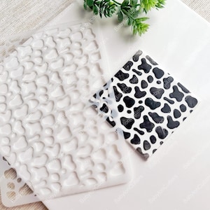 Op de afbeelding: Een witte plastic sjabloon met een patroon van onregelmatige, afgeronde vormen. De sjabloon is ontworpen om een ​​koeienprintpatroon te creëren. Een voorbeeld van het patroon wordt in zwart en wit getoond op een apart vel papier.