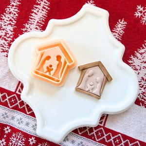 Op de afbeelding: Een koekjesvorm en een versierd koekje, beide met een kerststalscène in een huisvormig frame. De koekjesvorm is oranje en het koekje is bruin met bruine details. Op een wit, decoratief bord.
