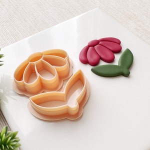 May include: Two plastic cookie cutters in the shape of a flower. The larger cutter is a daisy with five petals and a center circle. The smaller cutter is a leaf. The cutters are on a white surface.
