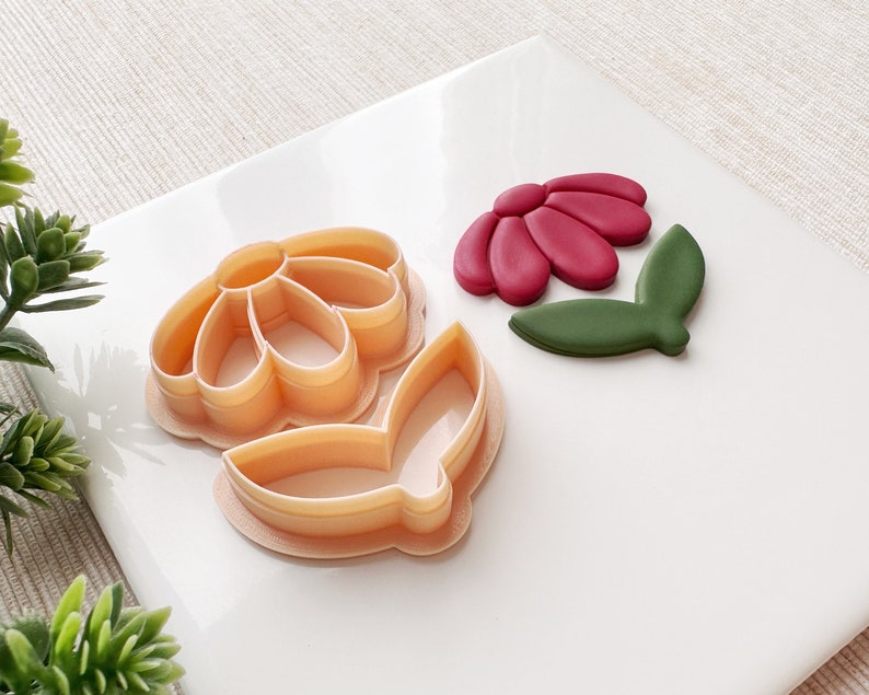 May include: Two plastic cookie cutters in the shape of a flower. The larger cutter is a daisy with five petals and a center circle. The smaller cutter is a leaf. The cutters are on a white surface.
