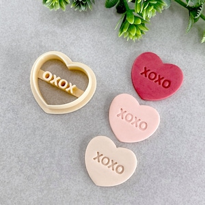 以下が含まれることがあります： 「XOXO」と刻印されたハート型のクッキー型が3つあります。1つは赤、1つはピンク、もう1つはベージュです。クッキー型は灰色の表面の上に置かれています。