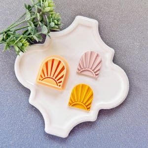 Op de afbeelding: Drie zonvormige koekjessnijders in oranje, roze en geel, gerangschikt op een witte, decoratieve schaal. De snijders hebben een sunburst-ontwerp in een boogvormig frame. De schaal staat op een grijze ondergrond, met een takje groen blad op de achtergrond.
