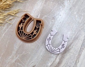 Puede incluir: Dos cortadores de galletas con forma de herradura con un diseño de sol naciente y detalles de cactus. El cortador de la izquierda es marrón y el de la derecha es blanco.