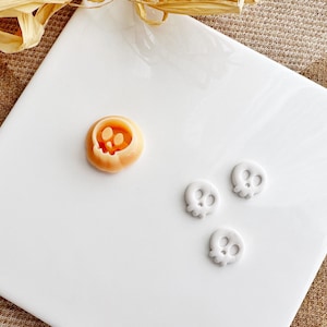 Op de afbeelding: Drie witte keramische knoppen in de vorm van een schedel op een wit oppervlak. Een van de knoppen is iets groter dan de andere twee. In de linkerbovenhoek van de afbeelding bevindt zich een kleine oranje keramische knop in de vorm van een schedel.