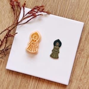 Puede incluir: Un cortador de galletas de plástico color melocotón claro con forma de figura estilizada con vestido largo y tocado puntiagudo, junto a una figura de arcilla verde oliva oscuro a juego con dos pequeños puntos rojos como ojos.
