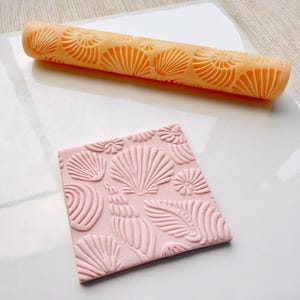 Op de afbeelding: Een close-up van een roze fondant met een schelpmotief, gemaakt met een deegroller met een schelpontwerp. De deegroller is oranje en heeft een reliëf schelpmotief.