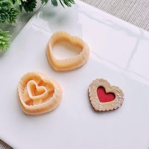 May include: Three heart-shaped cookies on a white surface. Two cookies are plain and one has a red heart in the center.