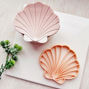 May include: Two pink and orange seashell shaped cookie cutters. The larger cutter is a solid pink color and the smaller cutter is a light orange color.