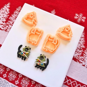 Puede incluir: Una placa cuadrada blanca muestra cortadores de galletas naranjas con forma de árboles de Navidad y gatos. También hay dos galletas decoradas con forma de gato. Las galletas son negras con decoraciones verdes en forma de árbol y luces de colores.