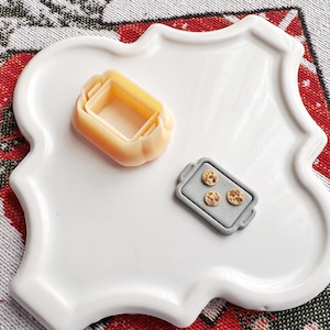Op de afbeelding: Een wit keramiek bord met een gegolfde rand heeft een miniatuur bakplaat met drie koekjes en een miniatuur bakvorm. De bakplaat is grijs en de bakvorm is beige.