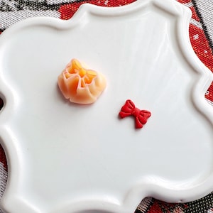 Könnte beinhalten: Zwei kleine Dekorationsartikel auf einem weißen Teller. Ein pfirsichfarbener, blumenförmiger Artikel und eine rote Schleife.