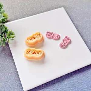 May include: Two pairs of miniature clogs in the image. One pair is a light peach color, and the other is a light pink color. The clogs are made of a rubbery material and have a detailed design. The clogs are on a white surface.