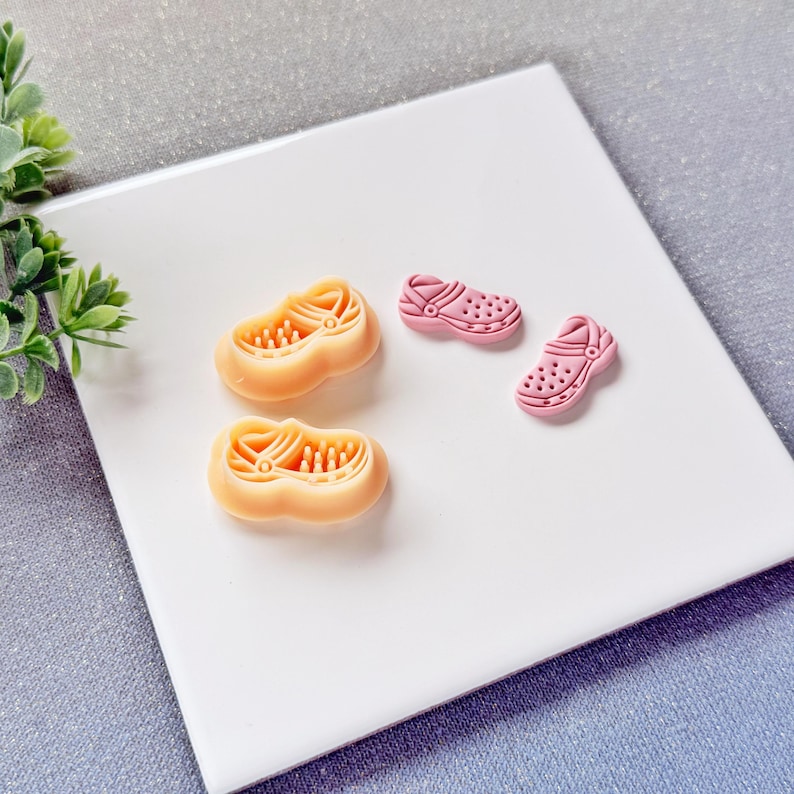 May include: Two pairs of miniature clogs in the image. One pair is a light peach color, and the other is a light pink color. The clogs are made of a rubbery material and have a detailed design. The clogs are on a white surface.