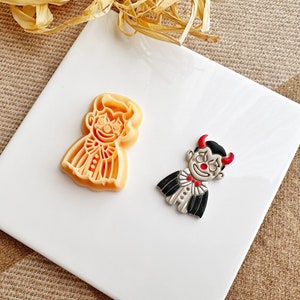 Op de afbeelding: Twee Halloween-thema koekjessnijders. Eén is lichtoranje en heeft de vorm van een persoon in een kostuum. De andere is een duivel met rode hoorns, een rode neus en een zwart-witte outfit. Beide liggen op een wit oppervlak.