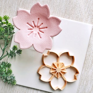 May include: A pink ceramic cherry blossom flower mold and a wooden cookie cutter in the shape of a cherry blossom flower.