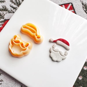 May include: Three Christmas-themed cookie cutters on a white surface. The cutters are shaped like Santa's hat, beard, and a hat outline. The hat and beard are colored red, white, and beige. The hat outline and beard outline are beige.