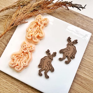 Pode incluir: Uma coleção de cortadores de biscoitos e biscoitos em forma de veado. Os cortadores são laranja claro, enquanto os biscoitos são marrons. Os veados têm chifres estilizados e um design antropomórfico único. Os itens estão dispostos em um prato quadrado branco.