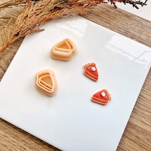 Può includere: Una collezione di formine per biscotti a forma di torta e fette di torta in miniatura. Le formine sono beige chiaro, le fette di torta rosse con crosta beige. Disposte su una superficie bianca con sfondo in legno.