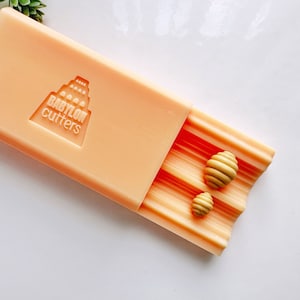 May include: An open peach-colored plastic case with the logo "BABYLON cutters" on the top. The case is open to reveal a set of small, yellow, honeycomb-shaped cookie cutters.