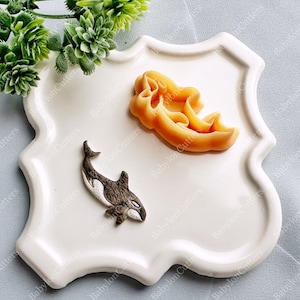 Op de afbeelding: Twee siliconen mallen voor het bakken, een in de vorm van een orka en de andere in de vorm van een werveling. De mallen liggen op een wit keramiek bord met een gegolfde rand.