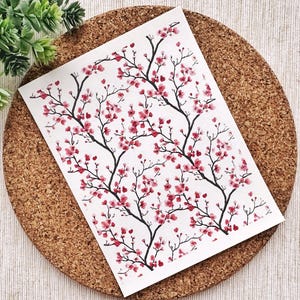 May include: A white card featuring a black and pink cherry blossom design. The card is displayed on a cork surface.