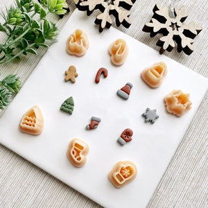 Könnte beinhalten: Ein Set mit 14 kleinen, bunten Ton-Anhängern in verschiedenen Weihnachtsformen, darunter ein Lebkuchenmann, ein Strumpf, ein Fäustling, eine Schneeflocke, ein Weihnachtsbaum, ein Zuckerstange, eine Glocke, ein Stern und eine Weihnachtsmannmütze.