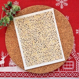 Op de afbeelding: Een rechthoekige kaart met een witte rand, met een beige achtergrond bezaaid met gouden en zwarte stippen. De kaart ligt op een ronde kurken onderzetter, geplaatst op een rood patroonweefsel met een takje groen gebladerte.