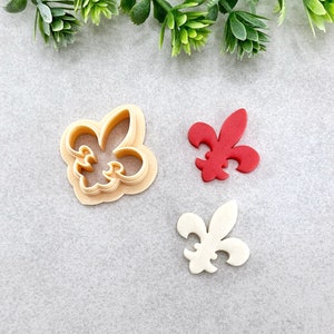 May include: Three cookie cutters in the shape of a fleur-de-lis. One is white, one is red, and one is a clear plastic cutter.