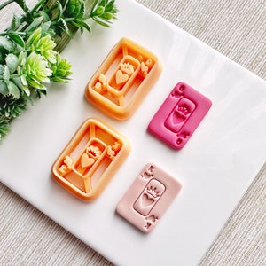 Op de afbeelding: Twee oranje plastic koekjessnijders in de vorm van speelkaarten en twee koekjes in de vorm van speelkaarten. De koekjes zijn roze en lichtroze, met een hart- en kroonontwerp. De koekjessnijders hebben een rechthoekig frame.