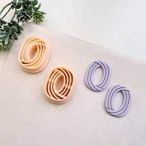 Pode incluir: Quatro brincos ovais de argila polimérica em tons de pêssego e lavanda. Dois brincos de pêssego têm um design em camadas, enquanto os brincos de lavanda apresentam um padrão tecido. Os brincos são exibidos em uma superfície cinza claro.