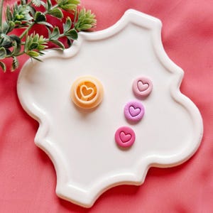 May include: A white, scalloped-edge ceramic plate holds a heart-shaped cookie cutter and three small, heart-shaped candies in shades of pink and purple. The cookie cutter is light orange. Greenery is in the upper left corner, and the background is pink.