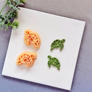 Pode incluir: Quatro moldes artesanais em forma de tartaruga. Dois são laranja com detalhes florais e dois são verdes com padrões florais. Os moldes estão em uma superfície branca, com folhagem verde no canto superior esquerdo.