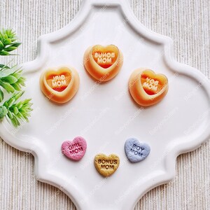 May include: Five heart-shaped candy molds with different colors and messages. The molds are on a white plate. The messages on the molds are "Girl Mom", "Bonus Mom", "Boy Mom", "I Love Mom", and "You Are Mom".